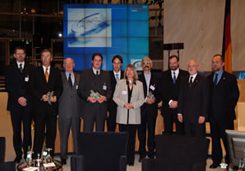 Mehrere Personen in Business-Kleidung stehen auf einer Bühne, hinter ihnen befindet sich ein großer eingeschalteter Monitor.